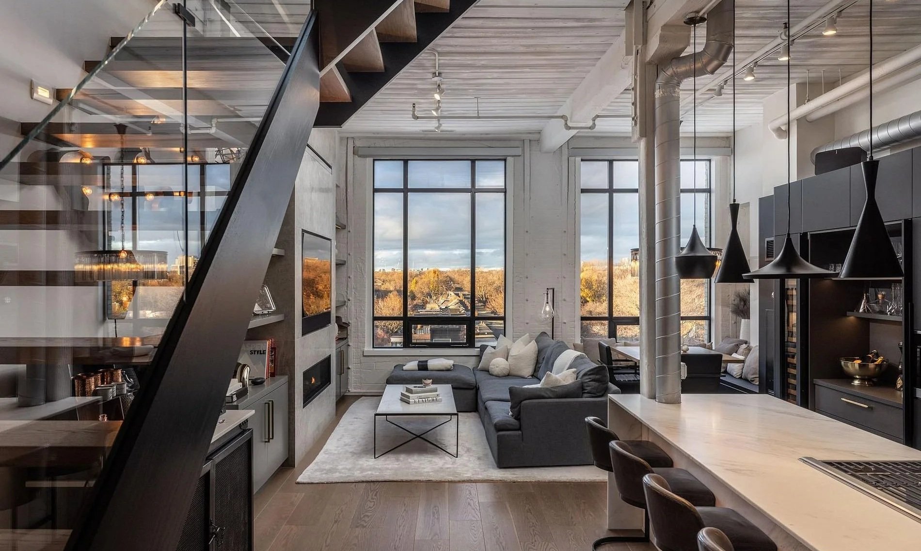 Modern living room with large windows showing cityscape and trees. Contains a sectional sofa with pillows, a coffee table, and a television. The room has an open ceiling with visible ductwork and black pendant lights hanging above the kitchen counter. The decor features neutral colors, and there is a staircase with glass railing visible on the left.